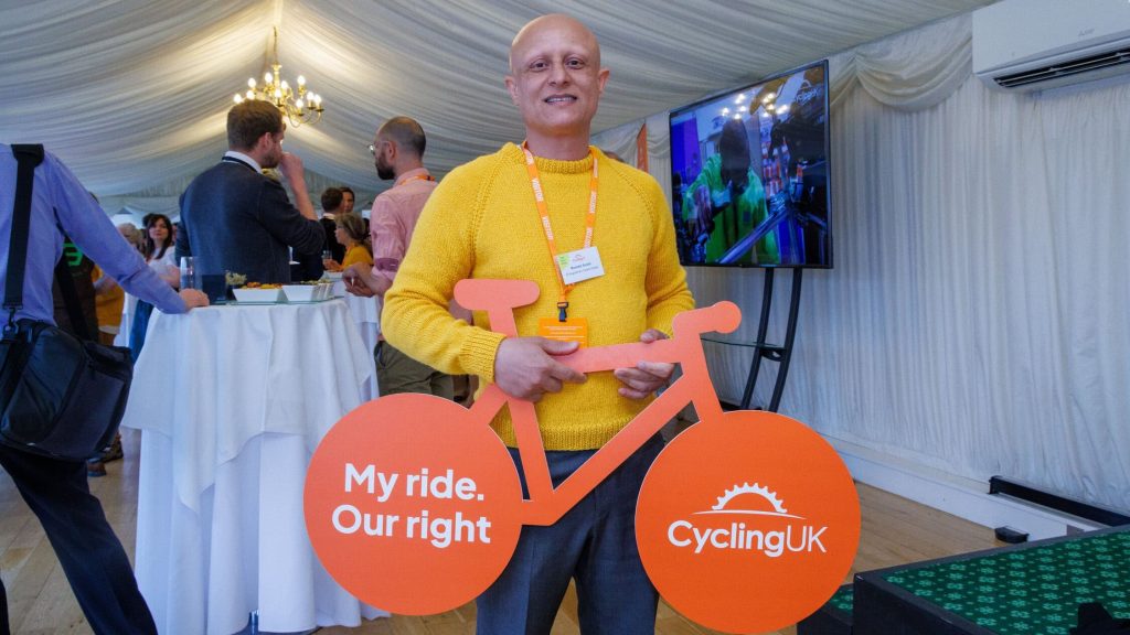 Person in a yellow sweater holding a large orange bicycle cutout with the text 'My ride. Our right' on the left wheel and 'CyclingUK' on the right wheel, standing in a room with other people and a screen in the background.