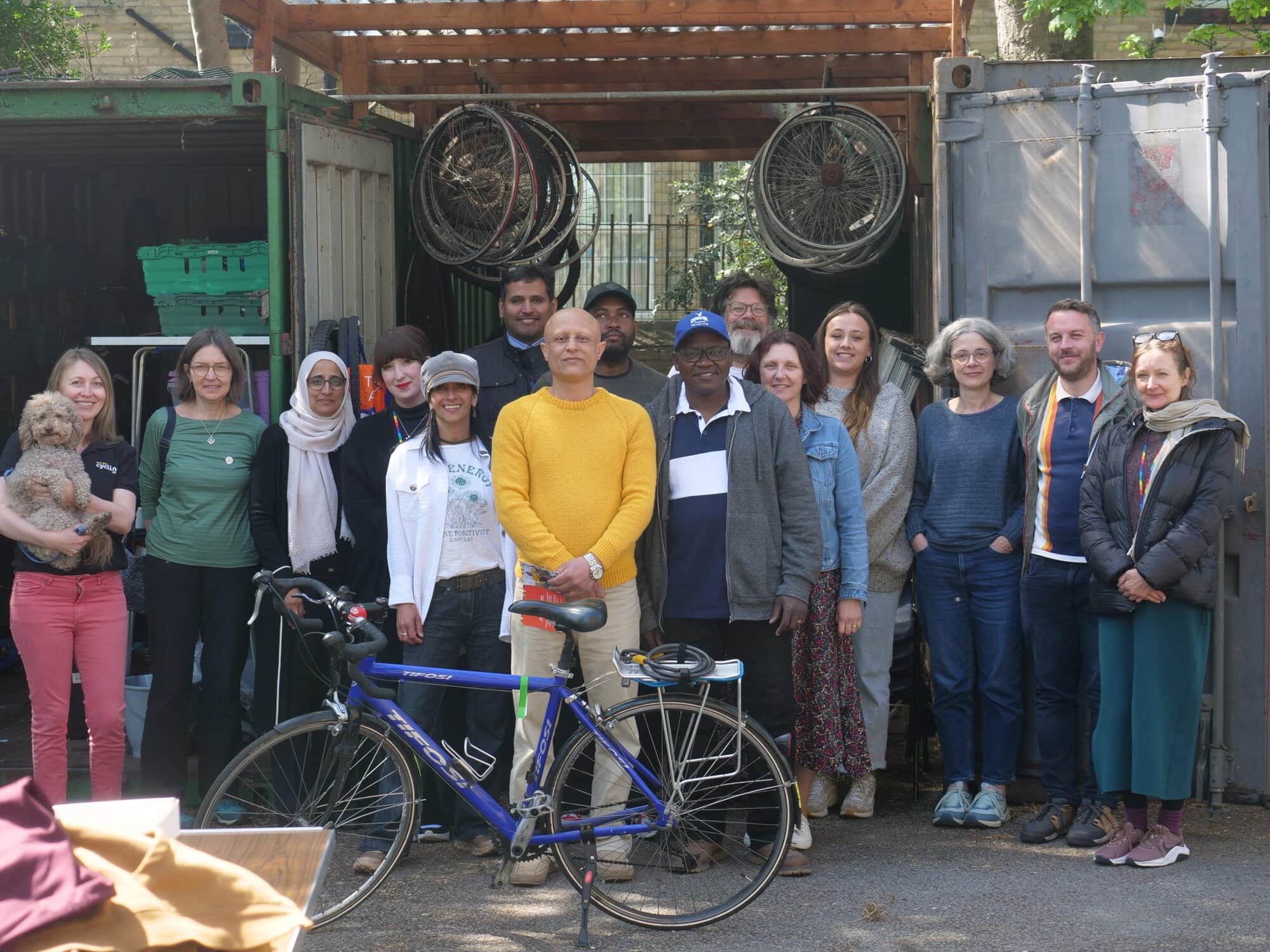 A group of people standing in front of a workshop area with two shipping containers and bicycle wheels hanging above. A blue bicycle is parked in front, and the faces are pixelated for privacy.
