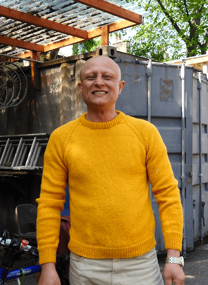 Mostafa in a bright yellow sweater and beige pants standing in front of a metal structure with ladders and bicycles in the background. Face is obscured by a blurred square.